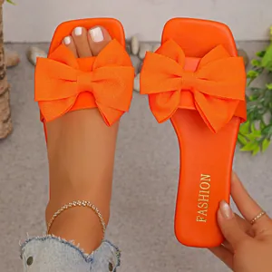 Detailed view of vibrant orange flat sandals featuring a large textured fabric bow on the strap.