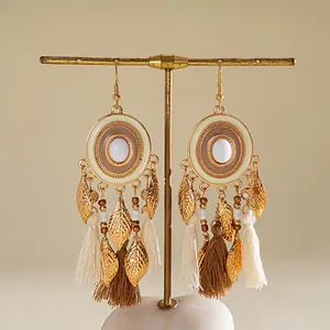 Gros plan détaillé de boucles d'oreilles bohèmes avec glands, feuilles d'or et résine blanche.