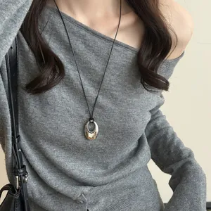 Detail eines ovalen Metallanhängers an einer schwarzen Lederschnur über grauem Pullover