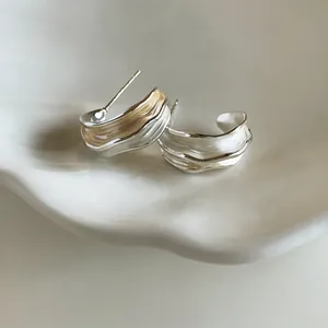 Détail de boucles d'oreilles argentées plissées au design ondulé sur fond blanc.