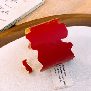Détail d'un bracelet et d'une bague en émail rouge avec des bords dorés irréguliers.