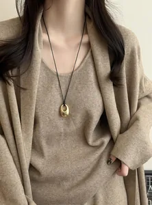Detailansicht einer goldenen ovalen Halskette mit schwarzer Kordel zu einem beigen Strickoutfit.