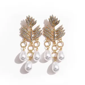 Elegante gouden blad oorbellen met strass en witte parel kwastjes op witte achtergrond.