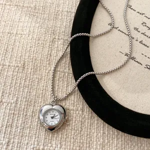 Détail d'un collier pendentif montre en forme de cœur argenté sur fond beige texturé.