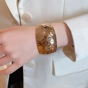 Close-up detail of a wide hammered gold-tone cuff bracelet on a wrist with white blazer