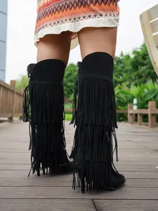 Detail view of black high-heeled boots featuring tiered long fringe on a wooden path