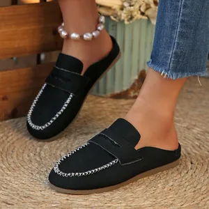 Black suede backless loafers featuring contrast white stitching on a straw mat background.
