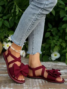 Nahaufnahme eleganter weinroter flacher Sandalen mit großer Schleife auf einem Holzboden.