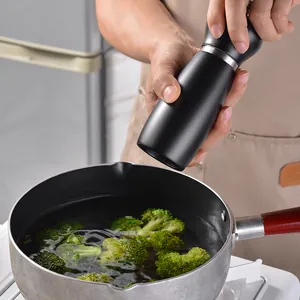Black manual pepper grinder in use over a cooking pot in a modern kitchen setting
