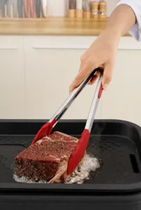 Red heat-resistant silicone food tongs used for searing a thick steak in a kitchen pan