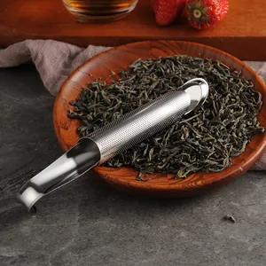 Pipe-shaped stainless steel tea infuser hanging on a wooden plate with tea leaves