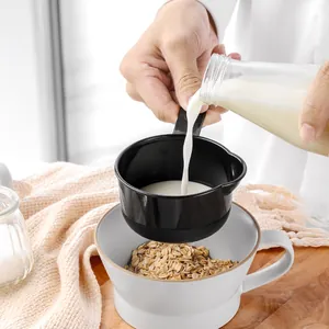 Black plastic measuring cup pouring milk into a bowl of oats for baking preparation.