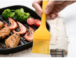 Yellow one-piece silicone BBQ oil brush held over a grill pan with cooked food