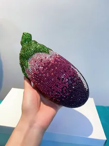 Hand hält eine einzigartige auberginenförmige Abendtasche mit lila und grünen Kristallen.