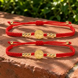 Par de pulseras de cordón trenzado rojo con medallas religiosas doradas sobre piedra.