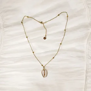 Detalle de un collar de acero dorado con colgante de caracola sobre tela de seda blanca.