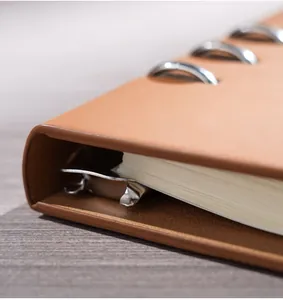 Primer plano de un cuaderno de anillas en cuero marrón con detalles de clips metálicos.