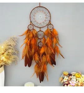 Λεπτομέρεια χειροποίητης ονειροπαγίδας boho με πορτοκαλί και καφέ φτερά σε γκρίζο τοίχο.