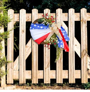 Corona patriótica del 4 de julio con bandera de EE. UU. y lazo de arpillera en valla.