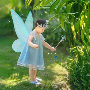 Bambina in costume da fata azzurro con ali, corona e bacchetta in un giardino lussureggiante.