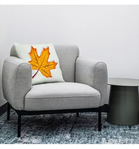 Square fall harvest pillow cover with an orange maple leaf design on a grey fabric armchair.