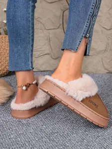 Close-up of tan fur-lined platform slippers worn with blue jeans on a grey rug.