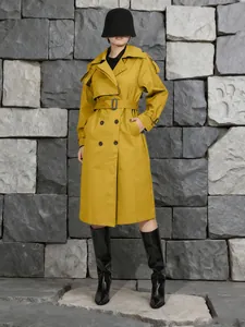 Elegante trench francese giallo senape con cintura davanti a un muro di pietra