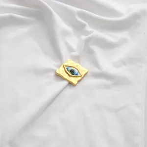 Vista detallada del adorno metálico dorado en forma de ojo en un top blanco sin mangas.