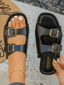 Detailansicht schwarzer offener Slipper mit zwei Bronzeschnallen an Frauenfüßen