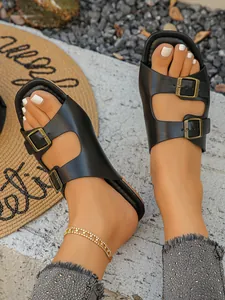 Nahaufnahme schwarzer Sandalen mit Doppelschnalle und goldenem Fußkettchen auf Stein