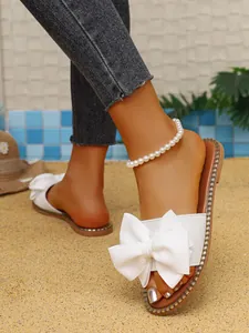 Detalle de sandalias planas blancas con un gran lazo decorativo sobre fondo de arena.
