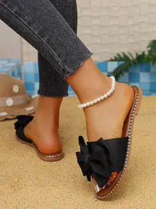 Detalle de sandalias planas negras con lazo y pulsera de perlas en fondo de playa.