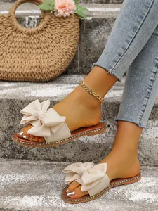 Detalle de sandalias planas beige con lazos grandes sobre escalones con un bolso de paja.