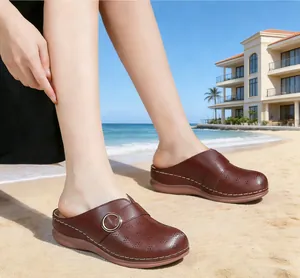 Side view of brown leather clogs with buckle detail worn on beach sand with sunny sky.