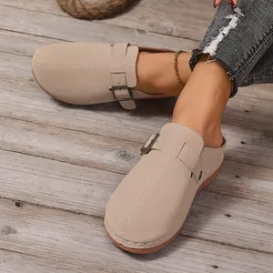 Elegant beige closed-toe mules featuring buckle strap detail on a rustic wooden floor
