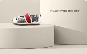Close-up of silver hollow-out baby leather shoes with red strap on a beige pedestal.