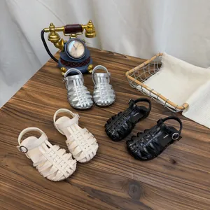 Gevlochten sandalen voor meisjes in zilver, zwart en beige op een houten tafelblad.