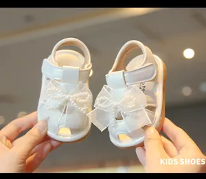 Elegant white baby girl sandals with lace bow and pearl details in close-up view.