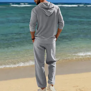 Rückansicht eines Mannes in einem grauen Leinen-Set mit Kapuze am sonnigen Strand.