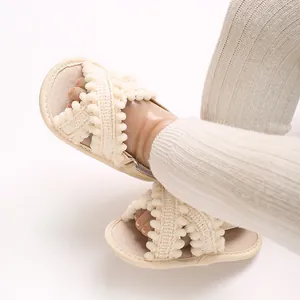 Primer plano de sandalias para bebé crema con correas cruzadas y detalles de pompones.