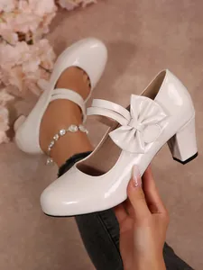 Dettaglio di scarpe Mary Jane bianche in vernice con tacco largo e fiocco decorativo.
