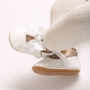 Close-up of white baby walker shoes with satin bow and sparkling rhinestone buckle detail.