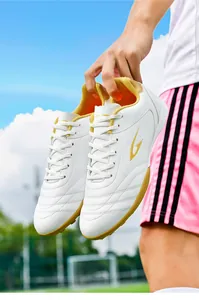 Close-up of white TF turf soccer shoes with yellow accents held in hand at a stadium.