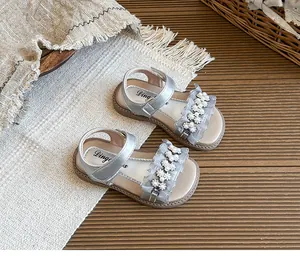 Top view of silver girls' sandals with pearl flower and ruffle details on a textured mat