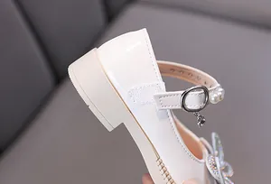 Primer plano de zapatos de charol crema para niña con lazo de pedrería y colgante de corazón.