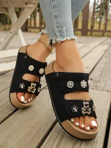 Nahaufnahme schwarzer Plateau-Sandalen mit Blumen- und Bären-Anhängern auf Holzdeck.