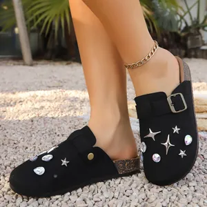 Close-up of black closed-toe mule slippers with sparkly rhinestone stars on outdoor gravel.