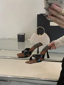 Λεπτομέρεια από μαύρα mules με ψηλό τακούνι και χιαστί λουράκια σε τραπέζι με καθρέφτη.
