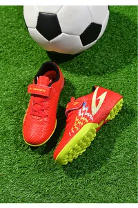 Dettaglio di scarpe da calcio rosse per bambini su erba sintetica con un pallone da calcio