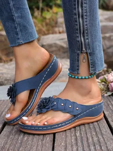 Detail van blauwe wedge teenslippers met decoratieve bloemen en klinknagels op houten vloer.